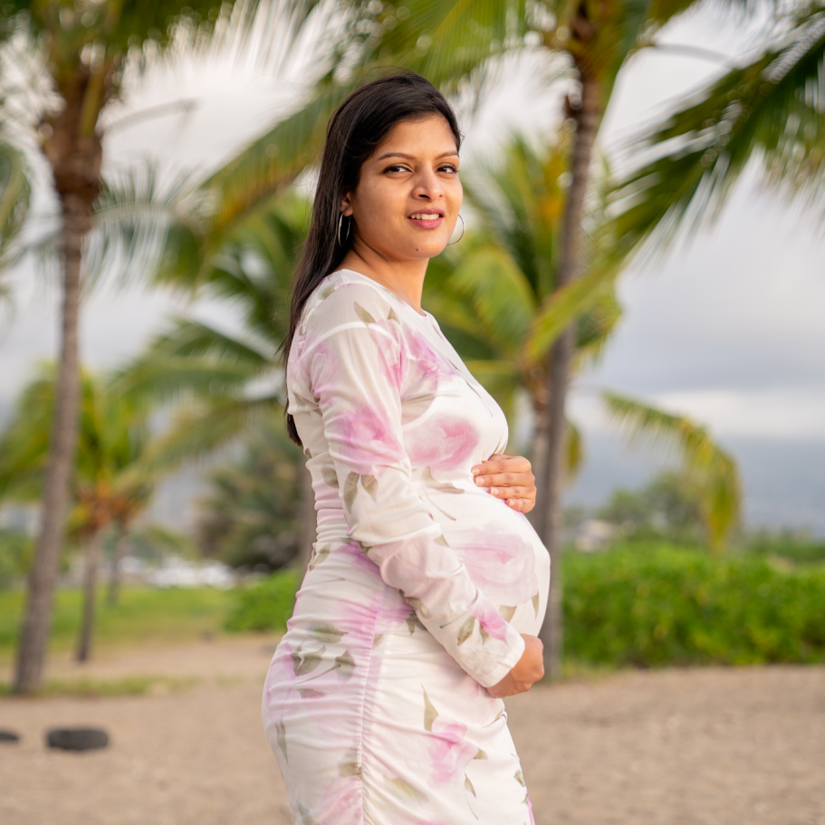 Pregnant woman in a floral dress standing on a beach with palm trees in the background big island photoshoot